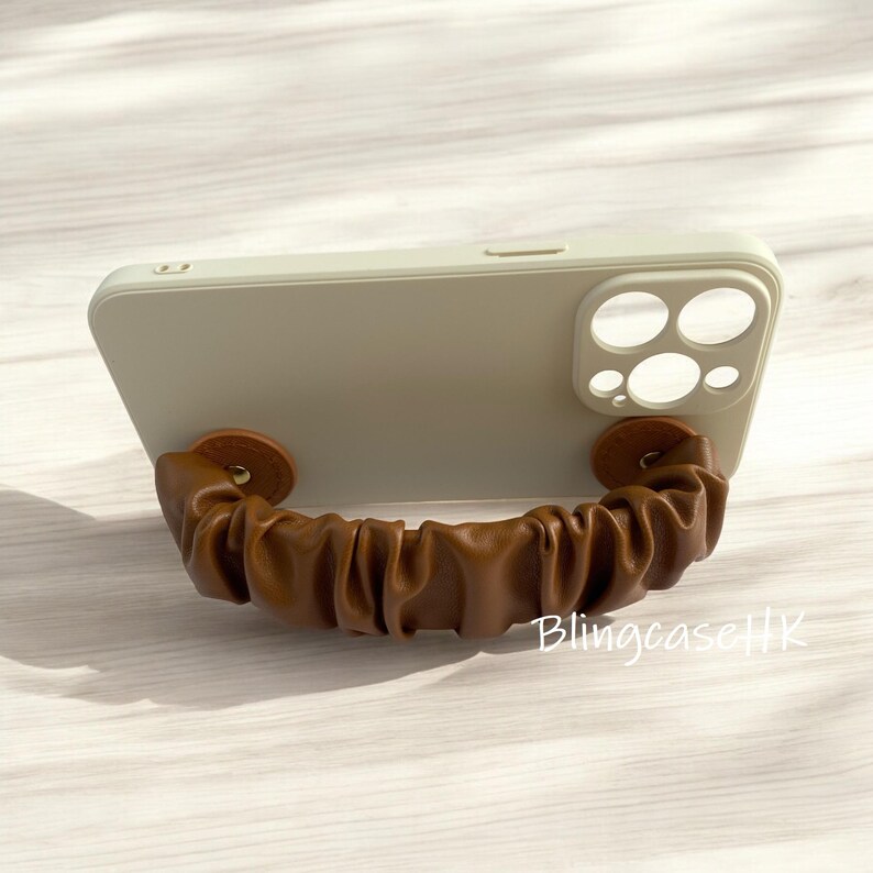 May include: A light green phone case with a brown leather strap attached. The strap is gathered and secured with two brown circular fasteners. The text "BlingcaseHK" is visible on the case.