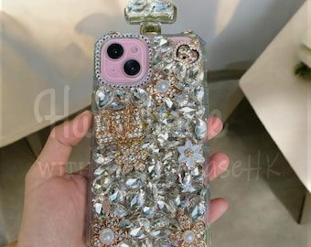 May include: A clear phone case with a pink interior, adorned with numerous sparkling rhinestones and faux pearls. The case features a gold-colored emblem and flower accents, with a crystal-like top.