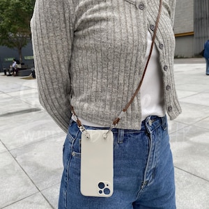 May include: A cream-colored phone case with a brown leather strap. The case is attached to the strap with silver-colored metal hardware. The phone case is being worn around the neck.