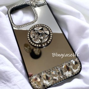 May include: A black phone case with a mirrored surface, embellished with sparkling rhinestones. The case features a ring holder and a decorative border of large, clear crystals. The text "BlingcaseHK" is visible on the case.