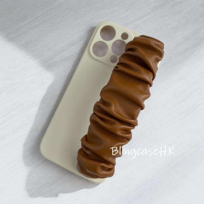 May include: A light beige phone case with a brown, ruched leather strap. The phone case has a camera cutout and is lying on a white surface. The text "BlingcaseHK" is visible on the strap.
