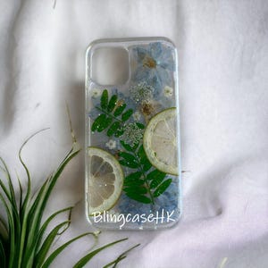 May include: Clear phone case with embedded dried flowers, green leaves, and lemon slices. The case features a transparent design with blue flowers and the text "BlingcaseHK".