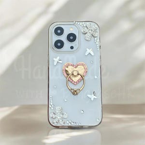 May include: Clear phone case with a decorative design. The case features a gold heart-shaped ring holder with pink accents, and is embellished with sparkling rhinestones in floral and star patterns. The case is designed to fit a smartphone.