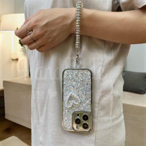 Puede incluir: Una funda de teléfono plateada con purpurina, diseño de corazón y marco dorado. La funda está sujeta a una correa de muñeca hecha de cuentas plateadas. La funda del teléfono tiene un recorte para la cámara.
