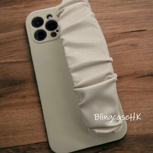 May include: A light beige phone case with a matching, ruched strap. The case has a glossy finish and a camera module with multiple lenses. The text "BlingcaseHK" is printed on the case.