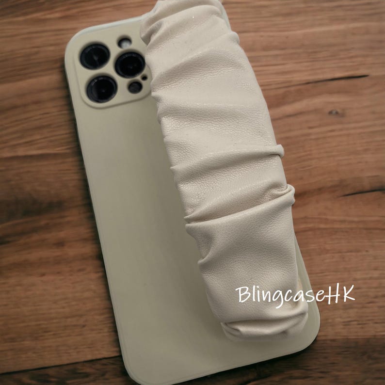 May include: A light beige phone case with a matching, ruched strap. The case has a glossy finish and a camera module with multiple lenses. The text "BlingcaseHK" is printed on the case.