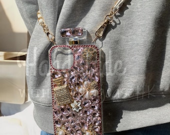 May include: A clear phone case with a pink crystal and pearl design. The case has a gold chain strap and a perfume bottle-shaped top. The case is decorated with pink crystals, gold accents, and white floral details.