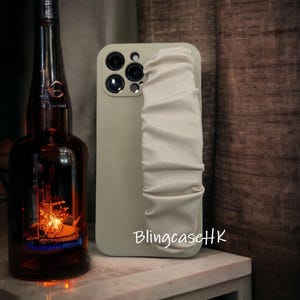 May include: A light green phone case with a white, ruched strap. The case has multiple camera lenses and the text "BlingcaseHK" in white. A dark brown bottle is in the background.