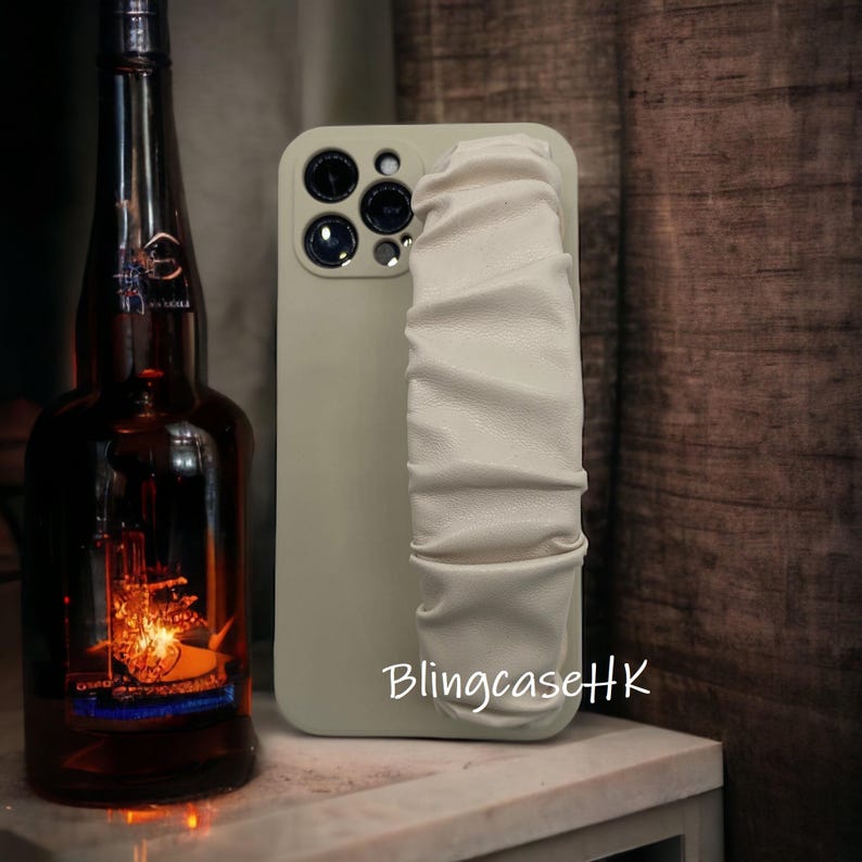 May include: A light green phone case with a white, ruched strap. The case has multiple camera lenses and the text "BlingcaseHK" in white. A dark brown bottle is in the background.