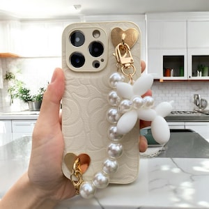 Puede incluir: Funda de teléfono color crema con un patrón floral en relieve, detalles dorados en forma de corazón y un dije de perro globo blanco y perlas. La funda tiene un cierre dorado y una correa de muñeca de perlas.