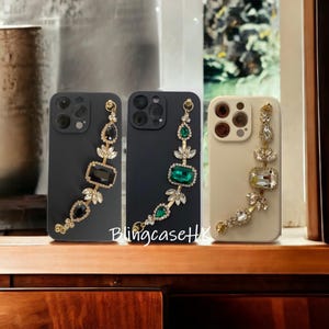 May include: Three phone cases with decorative jewel-toned accents. The cases are black and beige, with gold-colored chains adorned with black, green, and clear gemstones. The text "BlingcaseHK" is visible.