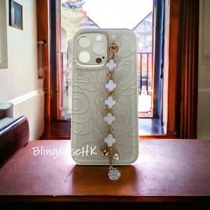 Op de afbeelding: Een lichtgekleurde telefoonhoes met een bloemenpatroon en een goudkleurige ketting met witte klavervormige bedels. De ketting heeft een parel en een bloemvormige bedel. De tekst "BlingcaseHK" is zichtbaar.