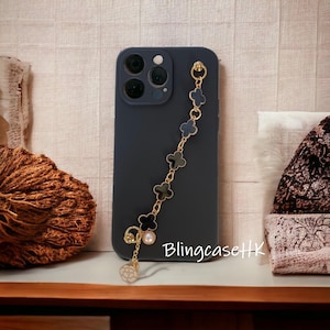 May include: A dark blue phone case with a gold chain featuring black and dark blue clover charms. The chain has a pearl and a white flower charm at the end. The text "BlingcaseHK" is visible on the case.