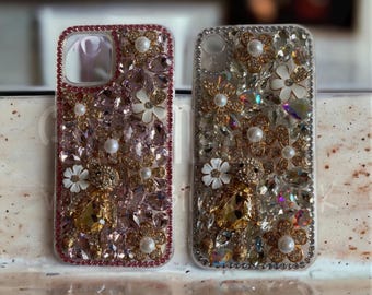 Op de afbeelding: Twee versierde telefoonhoesjes. Eén is roze met een rand, versierd met parels, witte bloemen en kristallen. De andere is zilver met vergelijkbare versieringen. Beide hoesjes hebben bloemen- en edelsteenontwerpen.