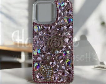 May include: A clear phone case bedazzled with pink and iridescent crystals. The case features a floral design with a pearl and gold accents. The case is designed to protect the phone while adding a touch of glamour.