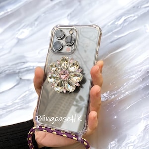 May include: Clear phone case with a large, sparkling flower design. The flower features clear and pink crystals. The case has a purple and gold lanyard. The text "BlingcaseHK" is visible on the case.