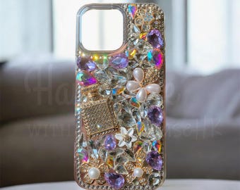 Op de afbeelding: Een telefoonhoesje versierd met een reeks heldere, iriserende en paarse kristallen, witte bloemvormige kralen en een goudkleurige bedel in de vorm van een parfumflesje. Het oppervlak is dicht bedekt met diverse fonkelende versieringen.