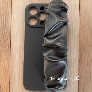 May include: Black phone case with a ruched leather strap. The case has cutouts for the camera and buttons. The text "BlingcaseHK" is visible on the bottom right corner. The case is on a wooden surface.