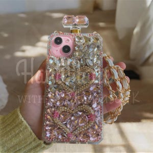 Puede incluir: Funda de teléfono adornada con cristales rosas y transparentes, con forma de frasco de perfume. Presenta una sección rosa para la cámara y una correa de cadena blanca y dorada. El texto "Ha with K" es visible.