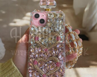 May include: A bedazzled phone case in the shape of a perfume bottle. The case is covered in pink and clear crystals, with a pink camera lens area. A white and gold chain strap is attached. The text "Ha with K" is visible.
