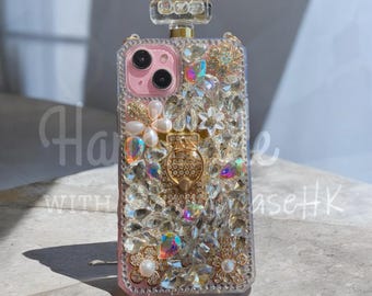 May include: A pink phone case with a perfume bottle design, encrusted with clear and iridescent rhinestones. The case features a gold-colored ring holder and is adorned with pearl and floral accents. The top has a clear, rectangular embellishment.