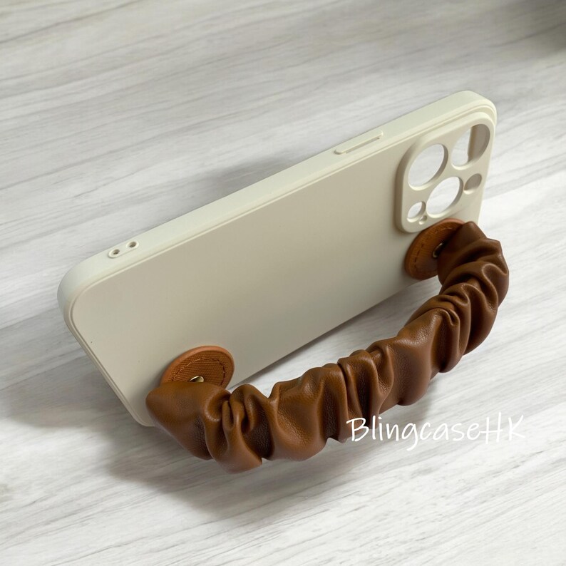 May include: A cream-colored phone case with a brown leather handle. The handle is attached to the case with two small, round, brown leather pieces. The text "BlingcaseHK" is visible on the case.