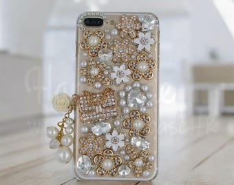 Op de afbeelding: Transparante telefoonhoes versierd met gouden, witte en heldere versieringen. Het ontwerp omvat goudomrande bloemen, parels en heldere kristallen. Een kleine gouden portemonneebedeltje en een witte rozenbedel zijn bevestigd.