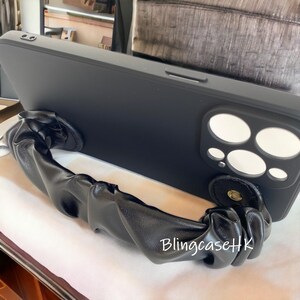 May include: Black phone case with a matching black, ruched wrist strap. The case has a camera cutout with white circles and the text "BlingcaseHK" in white.