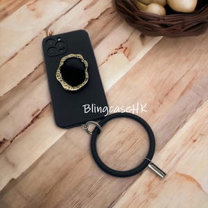 May include: Black smartphone case with a gold-colored, irregular-shaped ring holder. The case has a black wrist strap attached with a silver-colored clasp. The text "BlingcaseHK" is visible on the case.