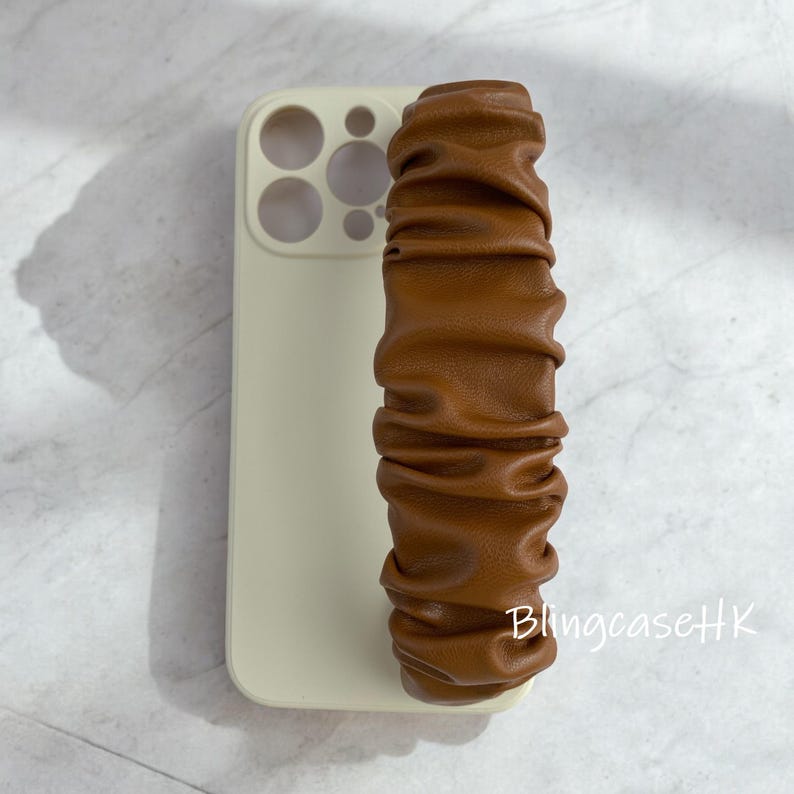 May include: A light beige phone case with a brown, ruched leather wrist strap. The strap is a long, rectangular shape with a gathered texture. The phone case has cutouts for the camera and buttons.
