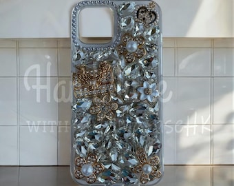 Op de afbeelding: Transparante telefoonhoes versierd met sprankelende strass-steentjes en goudkleurige bloemaccenten. De hoes heeft een kroon- en kettingontwerp. De hoes is versierd met een verscheidenheid aan heldere, facetgeslepen strass-steentjes.