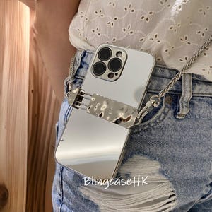 May include: A silver phone case with a chain strap. The case has a reflective surface and a textured metal accent. The chain is also silver and appears to be made of small, linked pieces. The text "BlingcaseHK" is visible on the case.