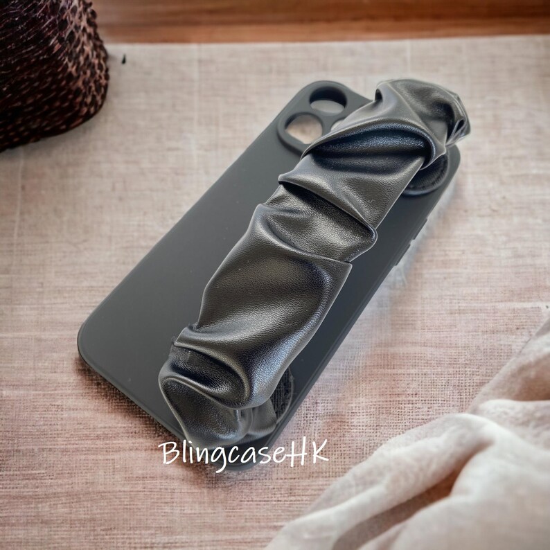 May include: A black phone case with a ruched leather strap. The case is matte black and has a camera cutout. The strap is gathered and adds a stylish element. The text "BlingcaseHK" is visible on the case.