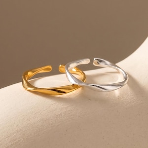 May include: Two open-ended rings with a smooth, undulating, twisted surface texture. One ring is finished in a polished gold tone, while the other ring is finished in a polished silver tone. Both rings feature a simple, continuous band design.