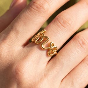 May include: A gold-toned ring featuring the word love written in a continuous, cursive script style. The metal band is formed by the letters of the word, creating an open, flowing design that wraps around the finger with a polished finish.
