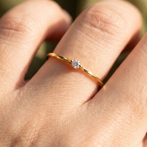 Peut inclure: Une fine bague en or ornée d'une petite pierre précieuse ronde et transparente sertie au centre. L'anneau présente un design délicat, légèrement torsadé ou texturé, ajoutant un détail subtil à la surface en or poli de la bague.
