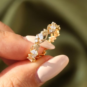 Könnte beinhalten: Ein goldfarbener Ring mit einem zarten, ineinander verschlungenen, rankenartigen Banddesign. Das Band ist mit kleinen, klaren, funkelnden Steinen besetzt, die in die Metallstruktur eingelassen sind und ein durchgehendes, filigranes Muster bilden.