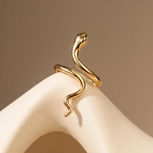 May include: A gold-toned ring shaped like a slender, winding snake. The metal has a smooth, polished finish, and the design features a continuous, curved body that wraps around to form an open-ended band with a defined head and tail.