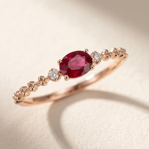 May include: A rose gold ring featuring a central oval-cut deep red ruby flanked by two small round white diamonds. The band has a delicate, textured beaded design that extends along the shank, creating a refined and detailed appearance.