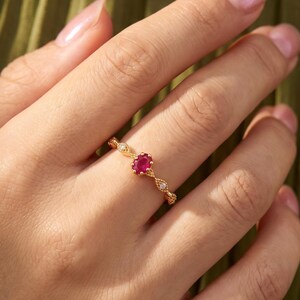 Könnte beinhalten: Ein Goldring mit einem zentralen, oval geschliffenen roten Edelstein, der von zwei kleineren, runden, klaren Steinen flankiert wird. Die Ringschiene weist ein filigranes, texturiertes Metalldesign mit feinen perlenartigen Details auf.