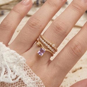 May include: A gold-toned ring set featuring a central rectangular light purple gemstone, accented by two additional bands with small clear stones. The rings are worn on a finger, showcasing a delicate, layered design with a polished metallic finish.
