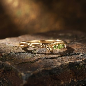 Puede incluir: Un anillo de oro con un diseño de banda trenzada que se asemeja a una enredadera o rama. El anillo está acentuado con tres pequeñas piedras preciosas de peridoto redondas de color verde vibrante y un pequeño diamante transparente en fila.