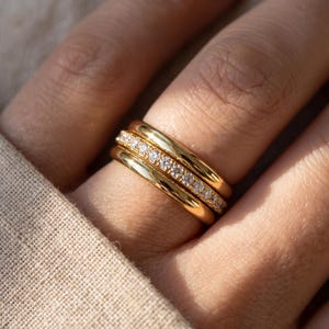 May include: A stack of three gold rings worn on a finger. The top and bottom rings are smooth, polished gold bands with a rounded profile. The middle ring is a gold band set with a single row of small, clear, round-cut stones that sparkle.