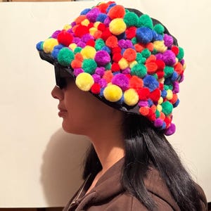 May include: A black hat covered in a variety of colorful pom-poms. The pom-poms are in shades of red, yellow, green, blue, and purple. The hat is worn by a person wearing sunglasses.