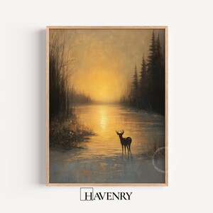 May include: A framed landscape print depicts a serene scene of a deer standing in a river at sunset. The artwork features a warm, golden sky, silhouetted trees, and a reflective body of water. The print is framed in a light-colored wooden frame.