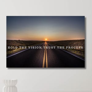 May include: A canvas print showing a road leading to a sunset. The image includes the text "HOLD THE VISION, TRUST THE PROCESS." The road is dark with yellow lines, and the sky changes from blue to orange. The print is displayed on a white wall.