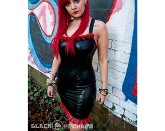 May include: A woman with long, bright red hair is wearing a black latex dress with a red ruffle trim. She is standing in front of a brick wall with graffiti.