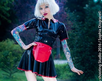 May include: A woman wearing a black latex dress with a red apron and black lace-up boots. She has white hair and is standing on a wooden deck.