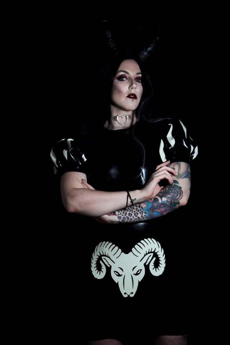 A person wearing a black latex dress with white accents and a heart-shaped choker. The dress has a white ram head design on the front. The person has black hair and is wearing black eyeliner and red lipstick.