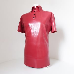 Può includere: Una polo a maniche corte realizzata in materiale lattice rosso lucido. La maglia presenta un colletto classico e un'abbottonatura frontale con quattro piccoli bottoni neri rotondi. Il materiale ha una superficie liscia e riflettente con pieghe visibili.